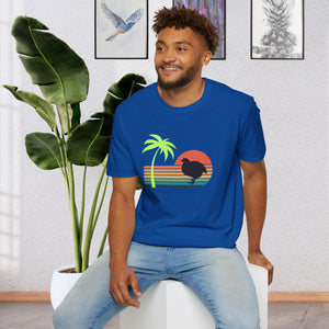 A man wearing a blue T-shirt that has a color line sunset ranging from orange at the top to teal at the bottom.  There is a coturnix quail in the middle of the sun and there is a vibrant green palm tree .