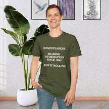 a man standing in a room in front of a plant wearing a Military Green T-shirt that says Homesteaders sharing information since 1862 Keep it rolling
