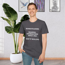 a man standing in a room in front of a plant wearing a Charcoal Grey T-shirt that says Homesteaders sharing information since 1862 Keep it rolling